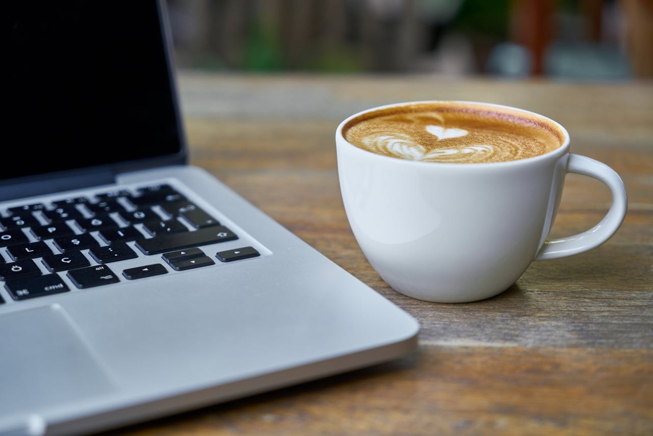 Services-03 Close-up of a latte coffee with heart art next to a laptop on a rustic wooden table.
