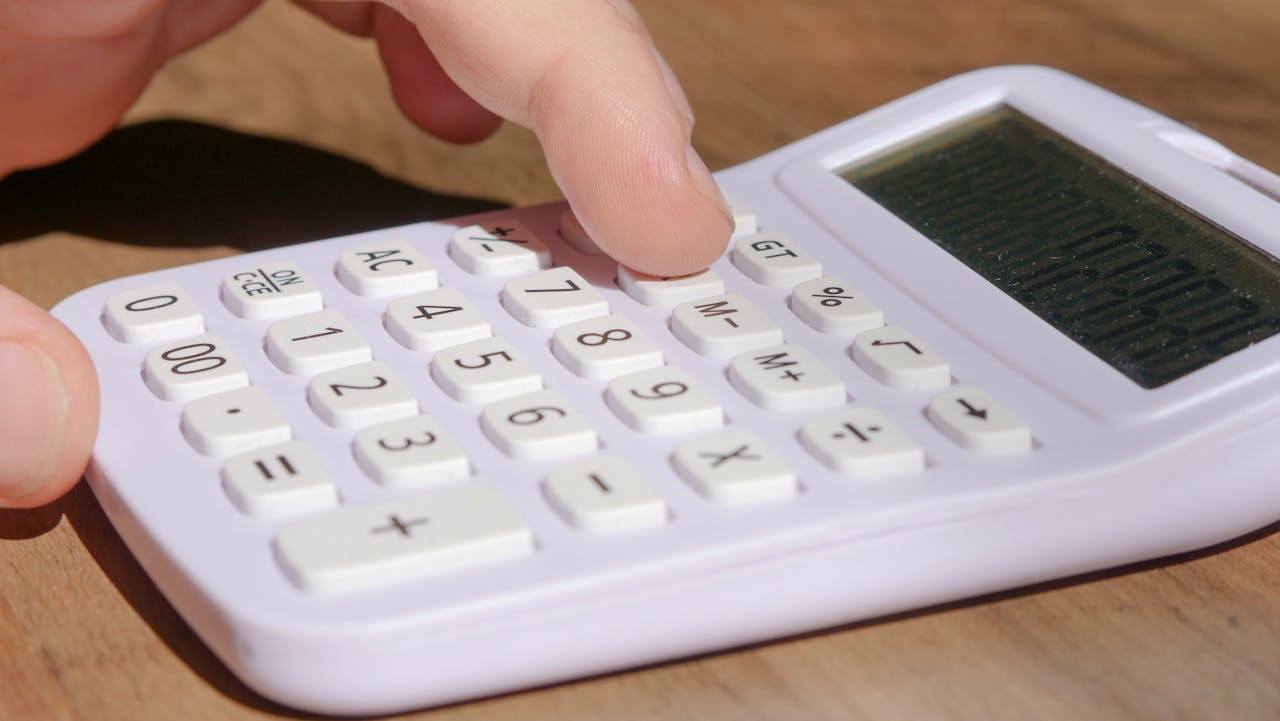 creative A hand pressing keys on a white calculator placed on a wooden desk in bright daylight.